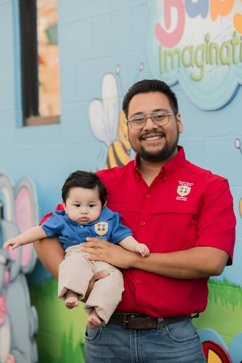 Man in red shirt holding smiling infant in blue outfit in front of colorful childrens mural