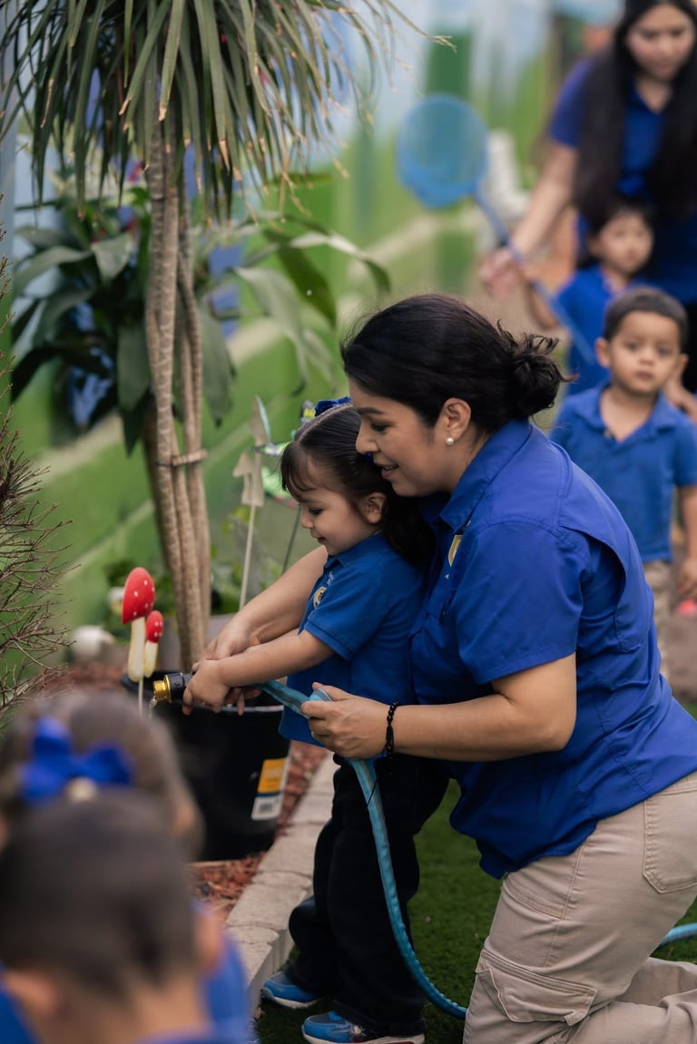 Woman in blue shirt helping young child water a red flower plant with a hose during outdoor group activity