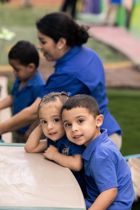 Two young children in blue shirts smiling at a table during an outdoor community event with adults supervising in the background
