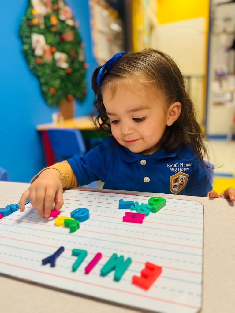 Young child in blue shirt arranging colorful magnetic blocks on lined paper at a classroom table with holiday decorations in background