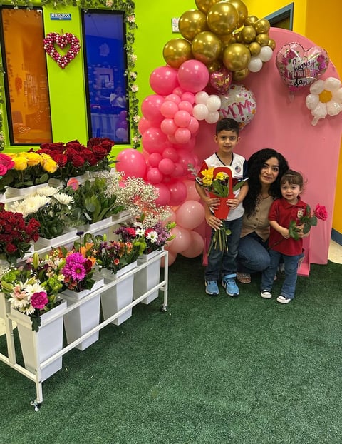 Woman and two children posing with flowers at a colorful Mothers Day photo display with balloons and floral decorations