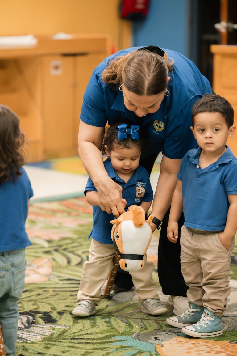 Teacher in blue shirt helps young toddlers interact with a toy horse during classroom learning activity