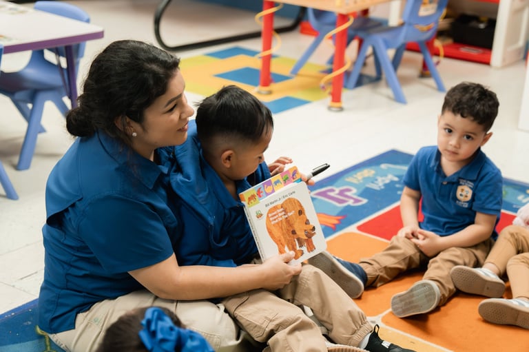 Teacher in blue uniform reads a picture book with two young boys seated on a colorful classroom floor with toys in the background