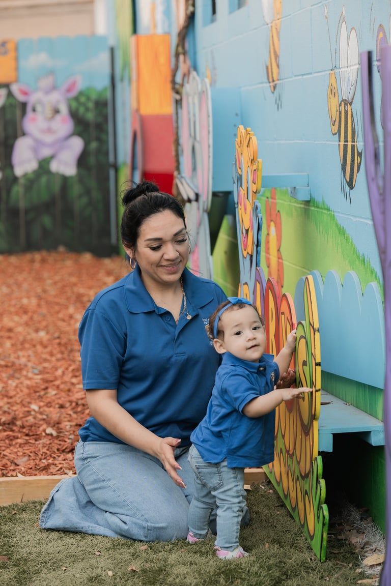 Woman in blue polo shirt kneels beside toddler wearing blue denim, both touching colorful mural with sunflower and animal designs
