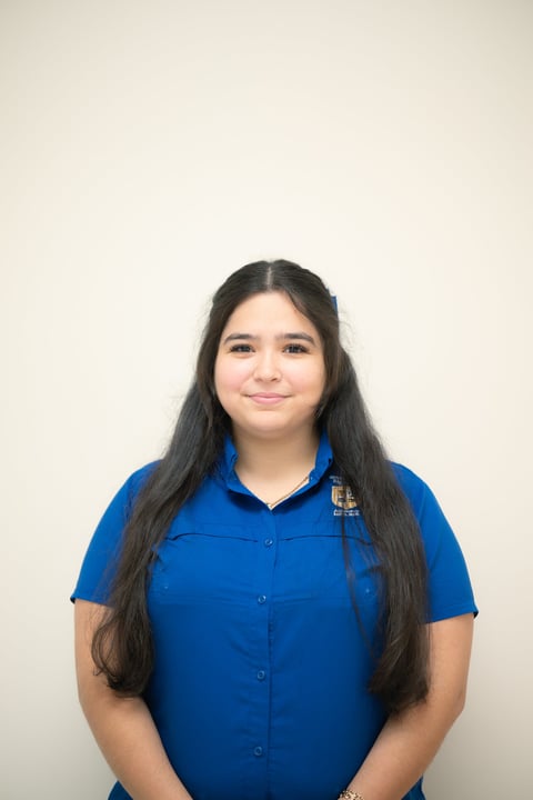 Portrait of a young woman with long dark hair wearing a blue button-up shirt against a neutral beige background