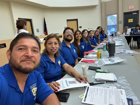 Group of people wearing blue shirts seated at a conference table with papers and materials during an official meeting or training event