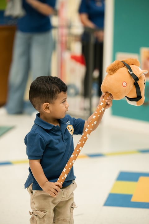 Young boy in blue polo shirt holding a tan hobby horse with patterned neck strap indoors