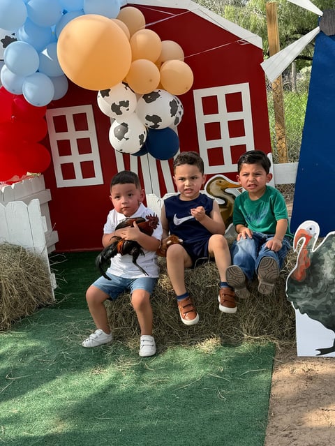 Three young children sitting together in front of a red playhouse decorated with balloons and cow print decorations