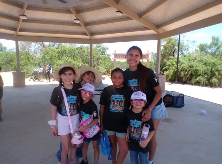 An adult woman with five young children wearing matching black camp t-shirts posing together under a covered pavilion outdoors
