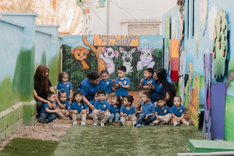 Group of young children and a teacher in blue shirts posing together in a colorful outdoor classroom with animal murals on the walls