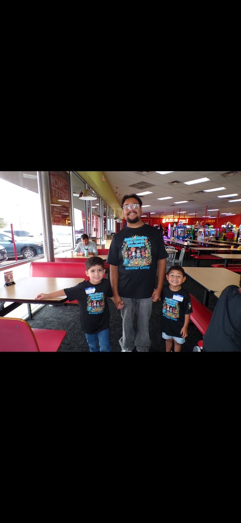 Adult and two children wearing matching black t-shirts standing in an arcade, with red seating and game machines visible