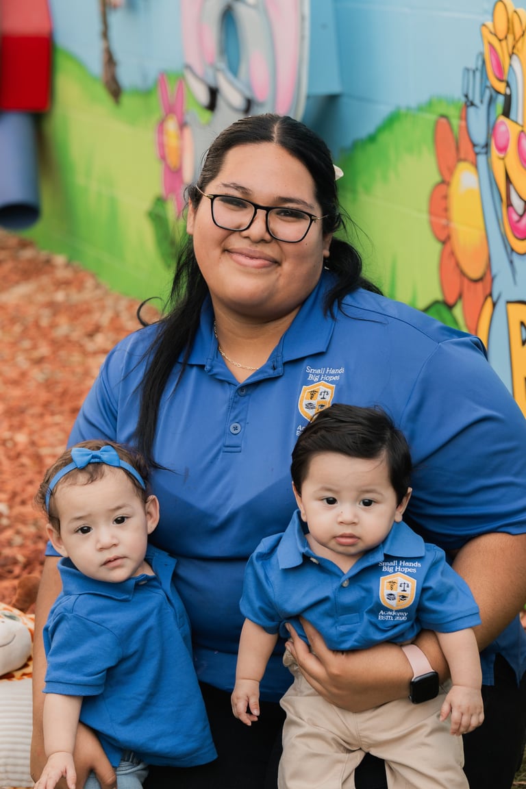 Woman wearing glasses and blue polo shirt with two toddlers in matching blue shirts against colorful mural background